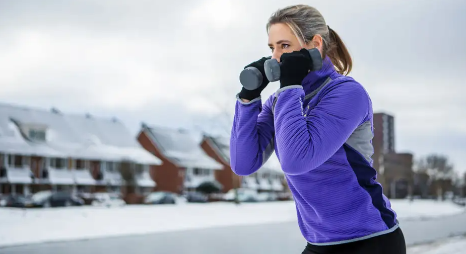 A person exercising outdoors in winter, holding dumbbells near their shoulders while wearing a purple jacket and gloves, with snow-covered houses in the background.