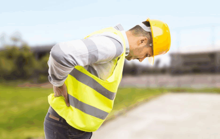“Construction worker wearing a hard hat and safety vest holding his lower back in pain at a job site.”